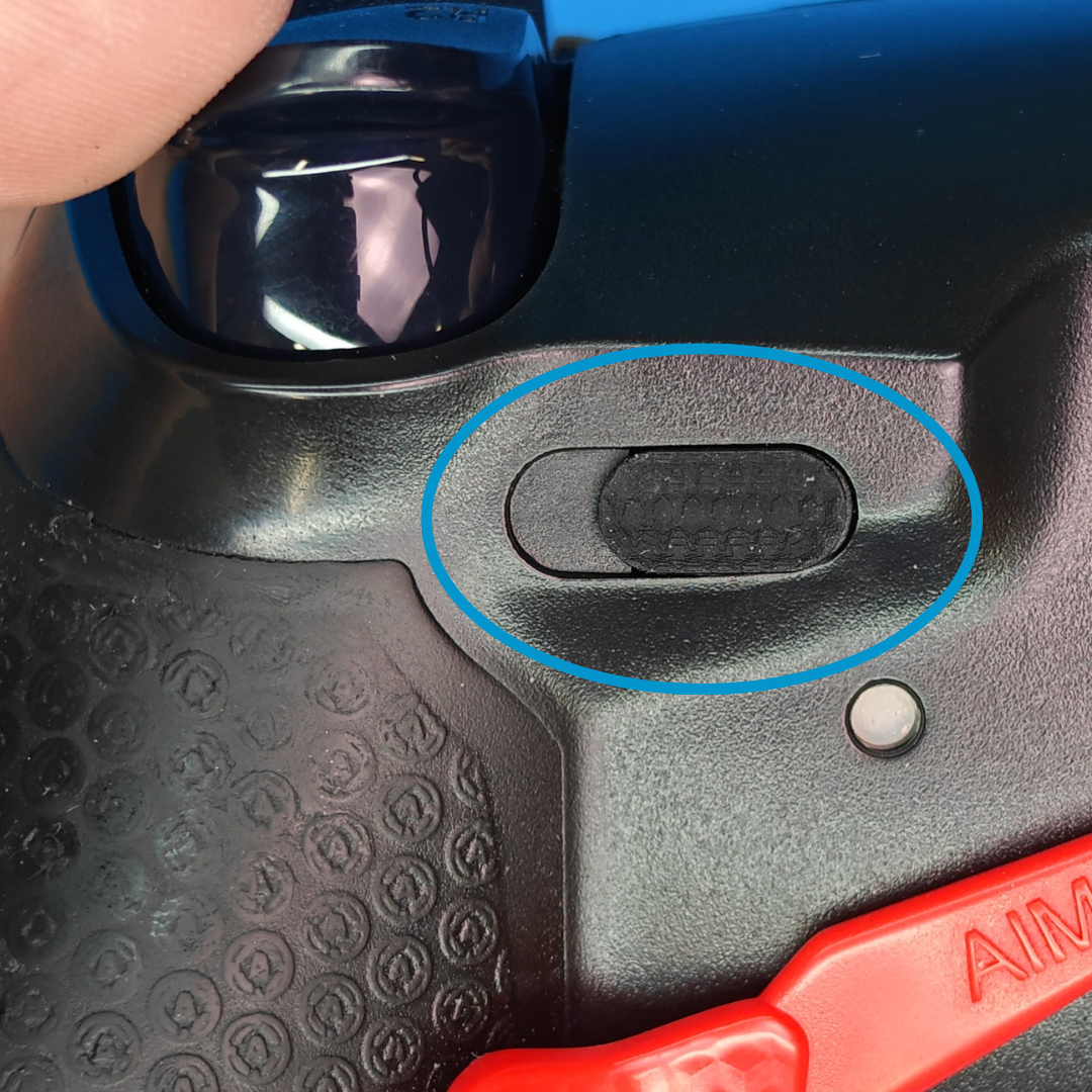 Close-up of the back of a black controller with a blue circle highlighting a small textured black button, with a red paddle visible below